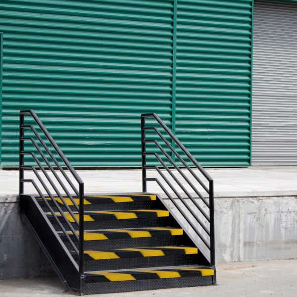 Black metal stairs with Anti-Slip Stair Nosing in yellow stripes lead to a loading bay, set before a green corrugated wall and closed grey rolling shutter door.