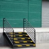 A small black metal staircase with yellow anti-slip stair nosing leads to a platform in front of a green corrugated metal wall and closed grey roller shutter door in an industrial or warehouse setting.