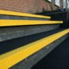 Concrete steps with Anti-Slip Stair Tread in bright yellow on each edge, beside a brick wall. The yellow treads enhance visibility and safety. Small plants and grass grow next to the wall.