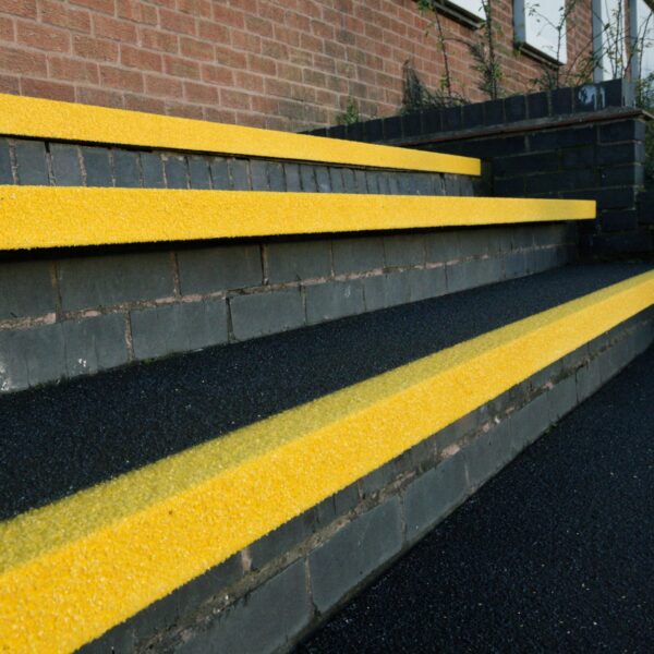 Concrete steps with Anti-Slip Stair Tread in bright yellow on each edge, beside a brick wall. The yellow treads enhance visibility and safety. Small plants and grass grow next to the wall.