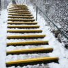 Outdoor concrete steps covered in snow feature bright yellow Anti-Slip Stair Treads on the edges. A metal handrail runs along one side, with a wire fence lining the path. Snow blankets the ground and surrounding areas.