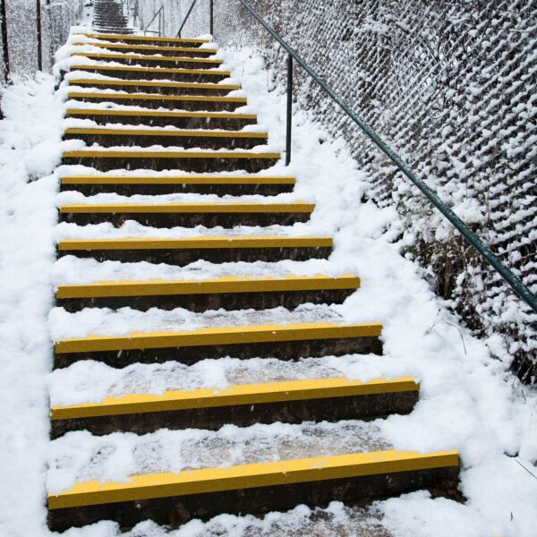 Outdoor concrete steps covered in snow feature bright yellow Anti-Slip Stair Treads on the edges. A metal handrail runs along one side, with a wire fence lining the path. Snow blankets the ground and surrounding areas.