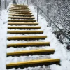 An outdoor staircase with Anti-Slip Stair Tread in yellow on each snow-covered step, bordered by a metal fence and handrail, on a winter’s day.