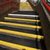 Outdoor stairs featuring Anti-Slip Stair Tread with black steps and yellow safety markings, bordered by brick walls and red railings, leading to a footbridge.