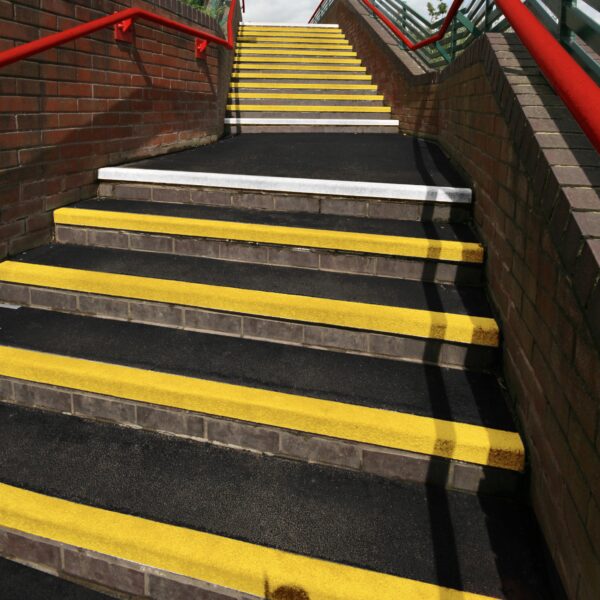 Outdoor stairs featuring Anti-Slip Stair Tread with black steps and yellow safety markings, bordered by brick walls and red railings, leading to a footbridge.