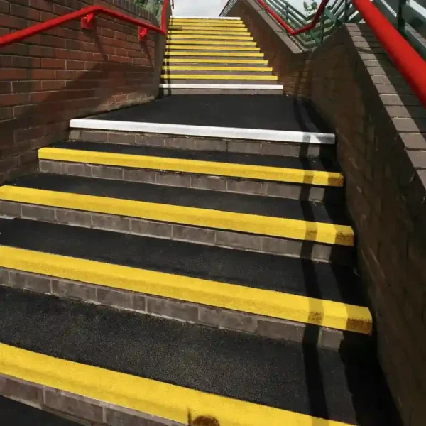 The Anti-Slip Stair Tread features black steps with bright yellow edges for visibility, red railings, and a brick wall topped with a metal fence on one side. One tread near the top is white.