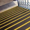 Dark brown stairs feature Anti-Slip Stair Tread with bright yellow safety strips on each edge, adjacent to a blue metal handrail and beige tiled upper floor.