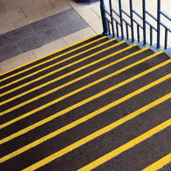 The Anti-Slip Stair Tread features dark steps with bright yellow safety lines on each edge beside a blue metal handrail, leading down to a tiled floor with a textured mat at the bottom.