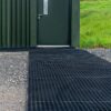 A dark green metal door with a small window sits in a corrugated wall. Leading to the door is a 16mm Black Grass Mat on gravel, with green grass on the right side.