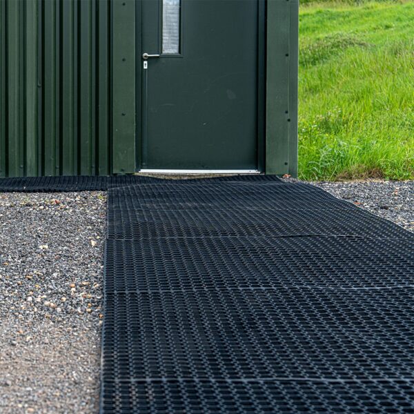 A dark green metal door with a small window sits in a corrugated wall. Leading to the door is a 16mm Black Grass Mat on gravel, with green grass on the right side.