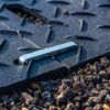 A close-up of the 2-Way Connector resting on a black textured panel with a diagonal slot, set on rough gravel. The connector aligns with the slot, indicating possible assembly or maintenance use.