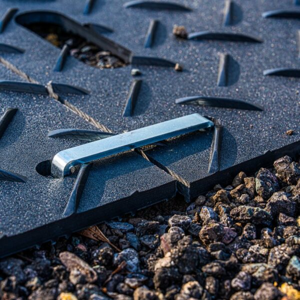 A close-up of the 2-Way Connector resting on a black textured panel with a diagonal slot, set on rough gravel. The connector aligns with the slot, indicating possible assembly or maintenance use.