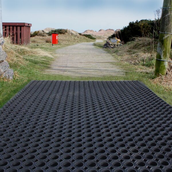 A Playground Safety Mat with circular holes sits on a wooden path among grassy dunes, near a red postbox and rustic fence, beneath a cloudy sky.