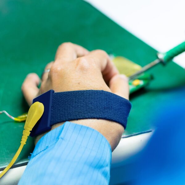 Someone wearing a blue Wristbands anti-static wrist strap, attached with a yellow wire, uses a soldering iron on an electronic circuit board placed on a green work surface.