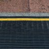 Close-up of a rooftop featuring brown asphalt shingles, a row of grey granules, a yellow cable, and the black ribbed Self-Draining Safety Mat below.