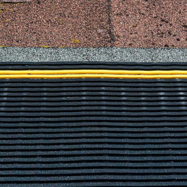 Close-up of a rooftop featuring brown asphalt shingles, a row of grey granules, a yellow cable, and the black ribbed Self-Draining Safety Mat below.
