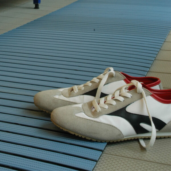 A pair of white trainers with black and grey accents and red interiors are displayed on a Cross-Ribbed PVC Leisure Mat placed atop a tiled floor.