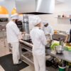A group of chefs in white uniforms and hats prepare food at stainless steel counters topped with bowls of ingredients and fresh vegetables, standing on a Nitrile Workplace Mat in a commercial kitchen.