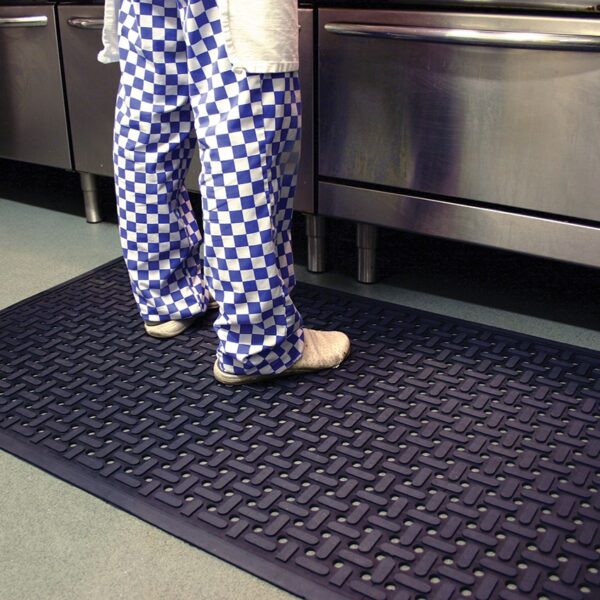 A person wearing blue and white chequered trousers and white shoes stands on a nitrile workplace mat in front of stainless steel kitchen cupboards, with only the lower half of the body visible.