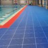 An indoor swimming pool features Multipurpose Interlocking PVC Tile in blue for non-slip flooring, with a narrow red strip along the edge. Flags, noticeboards, and equipment are visible in the background.