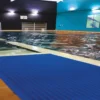 Indoor swimming pool with clear water, featuring a Leisure Mat in blue on the poolside. The area has blue and dark walls, bright ceiling lights, and a colourful artwork displayed on the right wall.