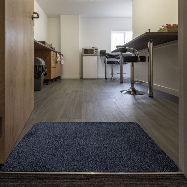 A modern kitchen with wooden cupboards, a microwave, and two bar stools at the counter. Sunlight enters through a small window, and an Internal Entrance Mat in blue is placed on the floor near the entrance.