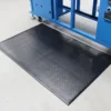 A Comfort Plus Anti-Fatigue Mat with a diamond plate texture in black is positioned on a light grey floor in front of a blue industrial machine.