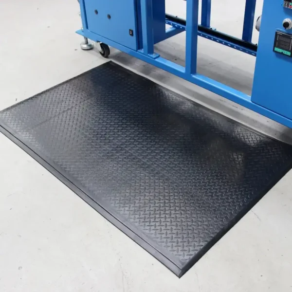 A Comfort Plus Anti-Fatigue Mat with a diamond plate texture in black is positioned on a light grey floor in front of a blue industrial machine.