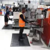 Two factory workers use industrial equipment; one operates a drill press in a high-visibility vest, while the other works in the background. Comfort Plus Anti-Fatigue Mats are placed on the floor for enhanced safety and comfort.