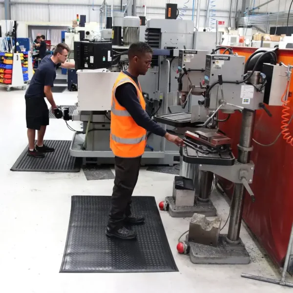 Two factory workers use industrial equipment; one operates a drill press in a high-visibility vest, while the other works in the background. Comfort Plus Anti-Fatigue Mats are placed on the floor for enhanced safety and comfort.
