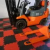 An orange forklift and pallet truck rest on a red and black Circular Stud Interlocking Tile floor beside a white brick wall.