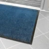 A Plush Entrance Mat with a blue rectangular surface and black rubber border is placed on a light grey tiled floor by a glass door with metal trim.