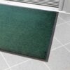 The Plush Entrance Mat in green with a black border is placed on a light grey tiled floor near a white sliding door.