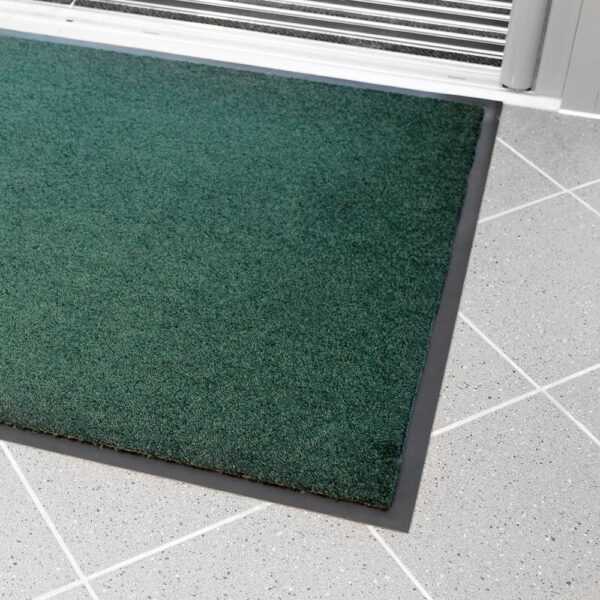 The Plush Entrance Mat in green with a black border is placed on a light grey tiled floor near a white sliding door.