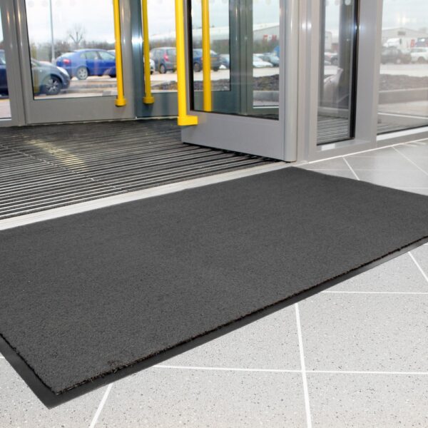 A Plush Entrance Mat in dark grey rests on a light grey tiled floor before a partially open glass entrance door, with cars visible outside through the glass.