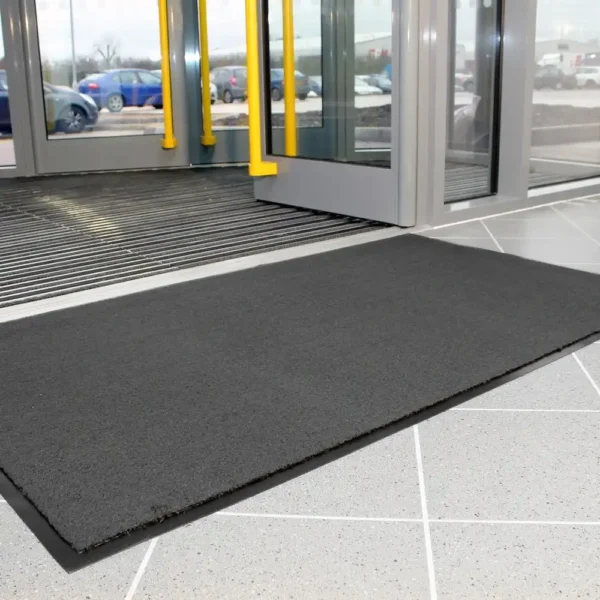 A Plush Entrance Mat in large black size is placed on a tiled floor by a glass door at a building entrance, with cars and trees visible outside through the windows.