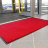 A Plush Entrance Mat in bright red is positioned on a tiled floor near the building’s glass doors, with parked cars visible outside.