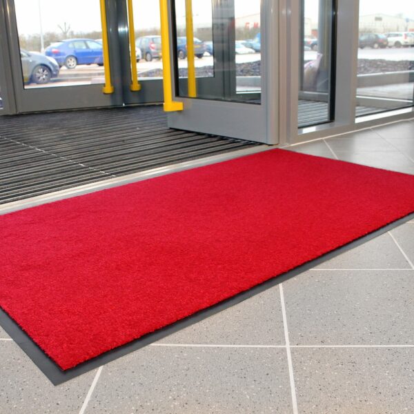 A Plush Entrance Mat in bright red is positioned on a tiled floor near the building’s glass doors, with parked cars visible outside.