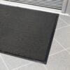 A plush entrance mat with a rectangular shape and black colour, featuring a rubber border, is placed on light grey tiled flooring near a metal-framed glass door.
