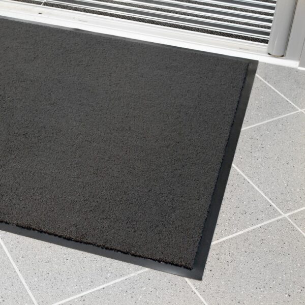 A plush entrance mat with a rectangular shape and black colour, featuring a rubber border, is placed on light grey tiled flooring near a metal-framed glass door.