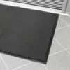 A close-up of the Plush Entrance Mat, a rectangular black doormat with a rubber border, placed on light grey tiles near a sliding glass door.