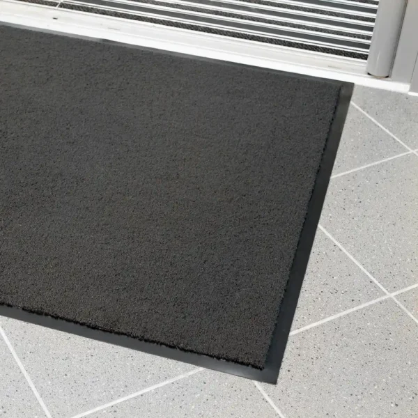 A close-up of the Plush Entrance Mat, a rectangular black doormat with a rubber border, placed on light grey tiles near a sliding glass door.
