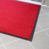 The Plush Entrance Mat, a bright red rectangle with a black border, is placed on a light grey tiled floor near a glass door.