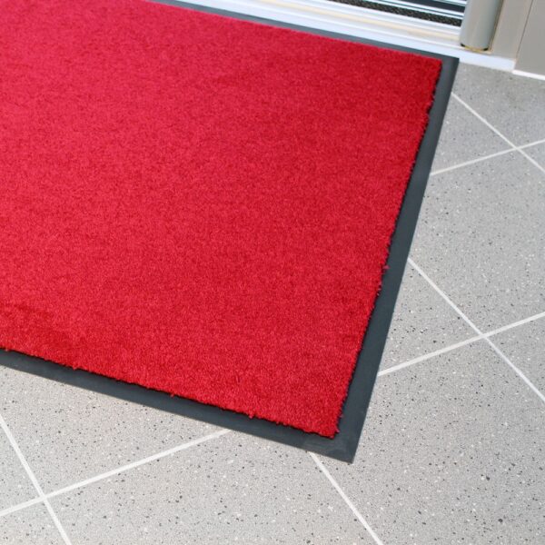 The Plush Entrance Mat, a bright red rectangle with a black border, is placed on a light grey tiled floor near a glass door.