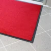 The Plush Entrance Mat, a vibrant red rectangle with a black border, is placed on a light grey tiled floor near a glass door.