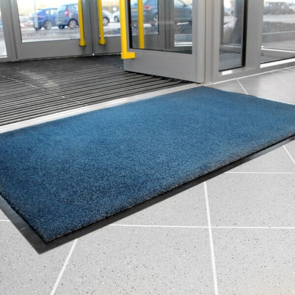 A Plush Entrance Mat is placed on a tiled floor by the building's glass doors with yellow handles, whilst cars can be seen outside.