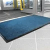 A Plush Entrance Mat in blue rests on a light grey tiled floor by glass doors with yellow handles at a building entrance; parked cars are visible outside.