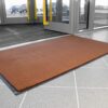 A Plush Entrance Mat in brown is placed on a grey tiled floor near large glass doors at a building entrance. Cars can be seen outside through the glass.