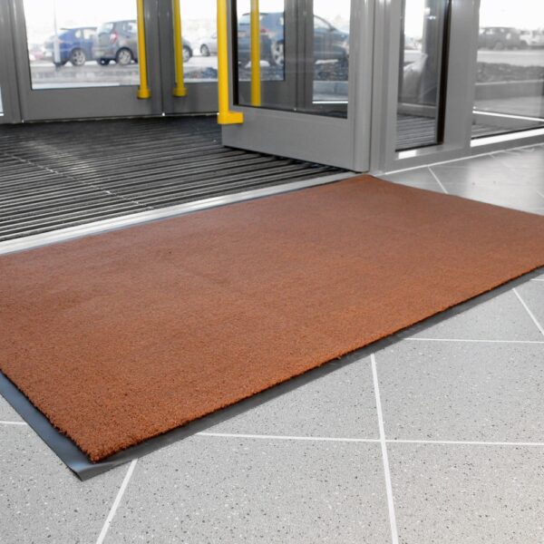 A Plush Entrance Mat in brown is placed on a grey tiled floor near large glass doors at a building entrance. Cars can be seen outside through the glass.