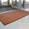 A Plush Entrance Mat in rectangular brown design is placed on a tiled floor near glass entrance doors, with cars visible outside.